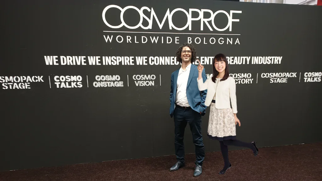 Trade show expert Ruben Uzan and Mary Condo posing in front of a sign saying Cosmoprof Bologna at the entrance of the trade show